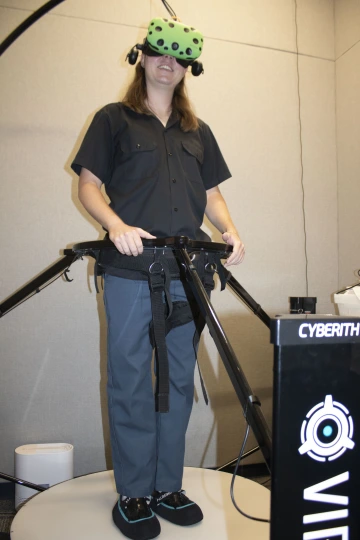 Josh stands strapped into a treadmill like machine with a VR headset on.