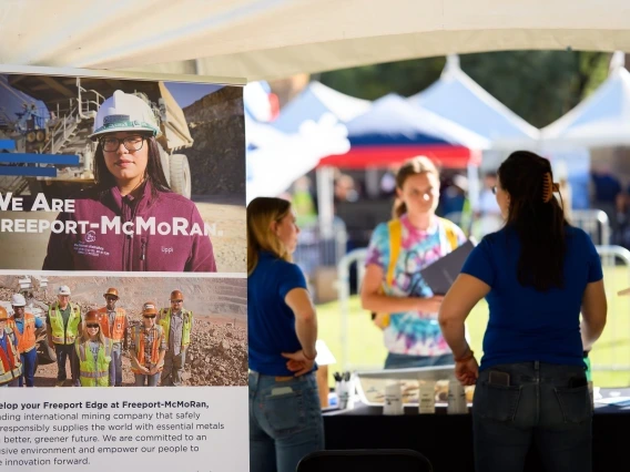 A booth display for Freeport-McMoRan at an outdoor event. A large sign features an image of a woman in safety gear with the text 'We Are Freeport-McMoRan,' alongside a group of miners in high-visibility clothing and hard hats. Below, text describes Freeport-McMoRan as an international mining company committed to safely supplying essential metals for a greener future. In the background, two people in casual attire engage in conversation near the booth, with other tents and event structures visible.