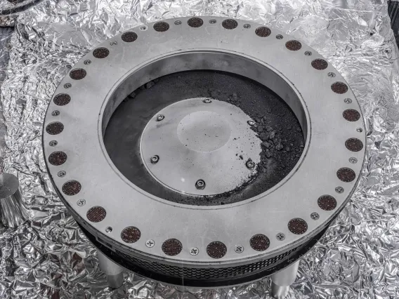 A round, metal canister with gray rocks and dust piled on the top