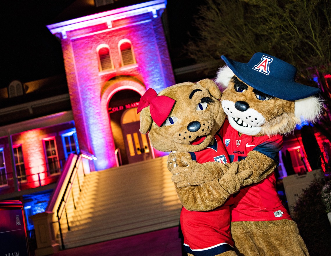 Wilbur and Wilma hug outside a blue and red lit Old Main. 