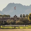 Wide view of the University of Arizona’s Old Main building in Tucson, Arizona, with the American and Arizona state flags flying above it, framed by tall palm trees and backed by mountains under a hazy sky. In the foreground, people walk dogs and take photos on the grassy mall, enjoying the sunny day.