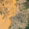 Satellite image showing a dense urban cityscape bordered by a large, muddy river with winding channels and sediment-rich water. The grid-like streets and clusters of buildings contrast with patches of green spaces, while bridges connect the city across the river.